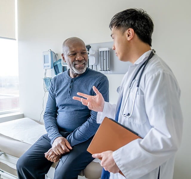 Doctor in white coat with stethoscope talking to smiling male patient seated in clinical examination room